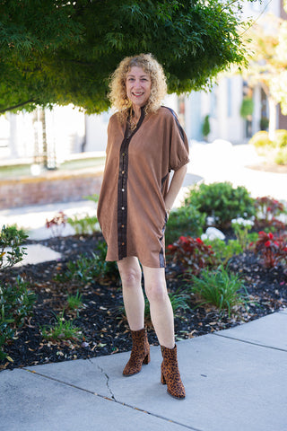 Woman posing outdoors on a sidewalk wearing a camel brown faux suede shift dress styled with leopard print ankle boots. The mini-length dress features a relaxed fit, short elasticized cuff sleeves, and contrasting dark brown faux leather trim down the central front placket with decorative metallic buttons. Styled for a fall fashion look with a chunky gold chain necklace under lush green trees.