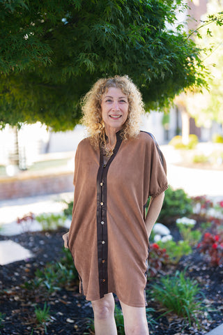 Medium shot of a smiling woman with curly blonde hair wearing a camel-colored faux suede tunic dress. The dress features a standing split collar lined with dark brown trim, metallic snap-front button details, short batwing-style sleeves, and side pockets. She has her hands tucked into her pockets, accessorized with a gold toggle chain necklace.