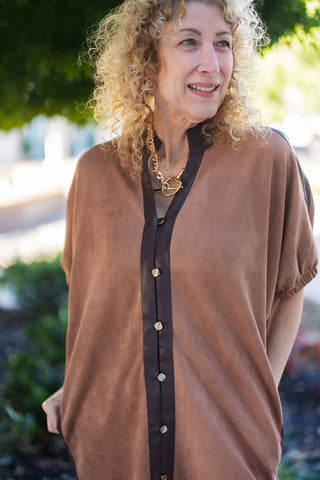 Close-up portrait shot emphasizing the texture and structural details of the faux suede dress. Highlights the dark brown contrast placket running down the center front with small square silver-toned metallic buttons. Showcases the V-neck split collar neckline and short sleeves with subtle elastic trim, worn with layered gold chain necklaces.