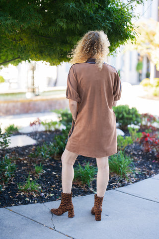 Back view of a woman wearing the camel faux suede mini shift dress paired with bold leopard print block-heel ankle boots. The shot shows the clean back silhouette of the tunic-style dress, short relaxed sleeves, and contrasting dark trim along the shoulder seams, set against a bright outdoor park backdrop