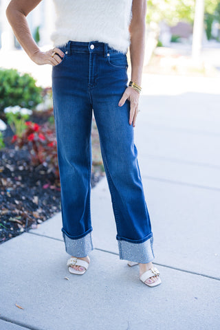Close-up view of the front waist and legs of dark wash high-waisted denim jeans, highlighting the structured belt loops, classic five-pocket styling, and the dazzling rhinestone-embellished turn-up cuffs paired with cream sandals.