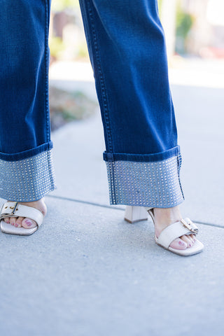 Detailed close-up on the hem of dark wash denim jeans, showcasing wide folded cuffs completely covered in shimmering rhinestones, styled with elegant white square-toe buckled block-heel sandals.
