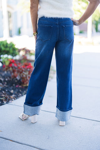 Back view of the dark wash straight-leg jeans featuring structured back patch pockets, a clean waistband fit, and the eye-catching crystal-embellished folded cuffs visible at the ankles above cream heels.