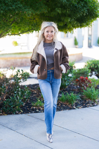 Woman posing outdoors on a sidewalk wearing a rich chocolate brown faux suede jacket lined with cream sherpa fleece. Features an oversized plush lapel collar, rolled sherpa-trimmed cuffs, large dark buttons, and welt hand pockets. Styled open over a charcoal graphic tee, high-waisted medium-wash blue jeans, and snakeskin patterned pointed-toe boots in front of landscaped greenery.