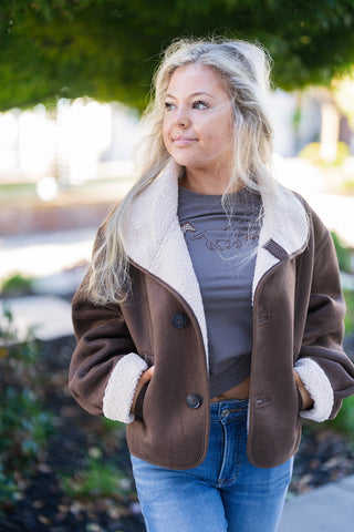 A portrait view highlighting the textured contrast of the dark brown suede fabric and soft cream faux shearling lining along the wide notch collar and folded cuffs. Styled open over a grey graphic top, displaying large dark front buttons, buttonhole stitching details, and side welt pockets with hands resting inside.