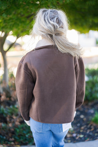 Back view of the rich brown suede jacket showcasing a boxy, slightly cropped silhouette with panel seam detailing across the back and shoulders. Highlights the cream sherpa collar peeking from behind and the clean hemline resting just above high-waisted denim jeans.