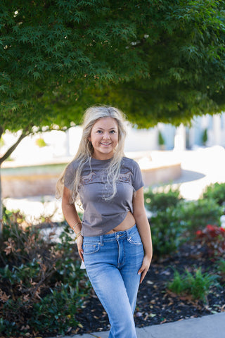 Full-length view of a smiling blonde woman wearing a fitted charcoal grey crewneck graphic tee featuring a horizontal line of running horses with gingham applique details. Paired with light-wash high-rise denim jeans and gold hoop earrings in a sunny outdoor setting.