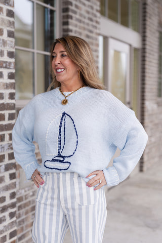A woman posing in a light blue crewneck sweater featuring a large, navy blue embroidered sailboat graphic on the chest. The sweater has a relaxed, slightly oversized fit with ribbed cuffs and hem, styled with gold coin and link necklaces. She is wearing high-waisted, light blue and white vertical striped trousers with front patch pockets, creating a cohesive maritime-inspired aesthetic against a neutral brick background.