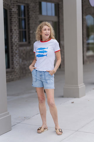 A woman stands on a sidewalk wearing the Dear John Julian Shorts in French Cloud, a soft, muted white denim. These mid-rise shorts feature a classic five-pocket design and a stylishly distressed fray hem for a relaxed coastal vibe. She has styled them with a white graphic tee with red trim and tan buckle slides.