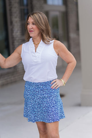 A portrait view of a woman wearing the Ditsy Daisy Skort, highlighting the high-definition miniature daisy pattern. The wide, smooth comfort waistband sits securely at the natural waist, paired with a crisp white ruffled-collar top and a gold cross necklace for a feminine, "Cottagecore meets Court-side" aesthetic.