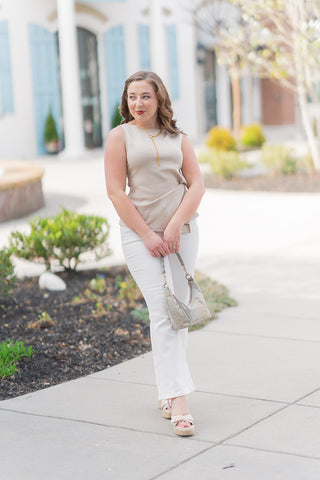A full-length front view of a woman wearing the Jayleen Knot Top, a sleeveless "elevated basic" in a refined sandy taupe neutral. The top features a high boat neckline and a sophisticated asymmetrical side-knot detail that creates a flattering drape across the bodice. Styled for a "quiet luxury" look, it is paired with high-waisted white denim, neutral platform espadrilles, and a gold lariat necklace, with the model holding a metallic snakeskin-textured handbag.