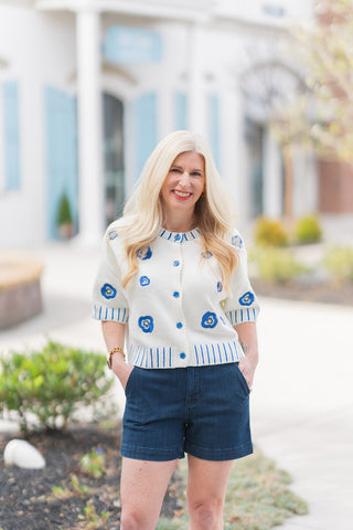 A full-length view of a woman posing in the Bluebell Bloom Top, a cream knit short-sleeve cardigan featuring vibrant blue floral embroidery. The top is styled with high-waisted dark indigo denim shorts, showcasing the flattering cropped hemline that hits at the waist. The model stands in a bright outdoor plaza with a background of classic architecture and light blue shutters, emphasizing a fresh, seasonal spring look.