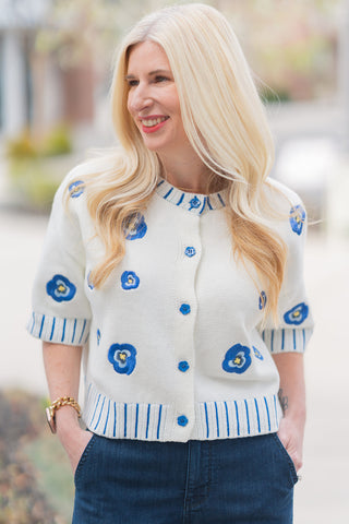 A portrait view highlighting the artisanal details of the Bluebell Bloom Top. The image showcases the intricate multi-toned blue floral motifs scattered across the cream knit and the tonal blue buttons on the front placket. Details like the blue vertical contrast stitching on the round neckline and the wide ribbed hem are visible, offering a high-quality, boutique aesthetic. The model’s blonde hair is styled in loose waves, complementing the top's romantic, whimsical feel.