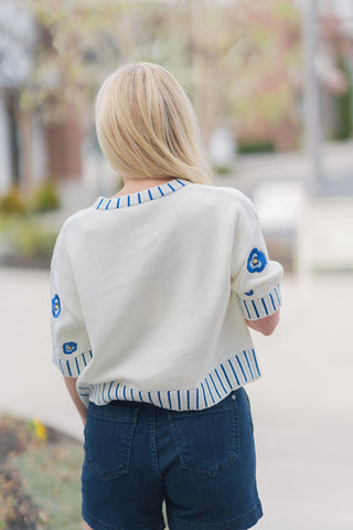 The back view of the Bluebell Bloom Top highlighting the relaxed fit and drop-shoulder design. The blue floral embroidery continues across the back and sleeves, while the decorative blue vertical stitching frames the neckline, cuffs, and hem. Paired with dark denim, this view emphasizes the clean, cohesive design and the lightweight, breathable texture of the knit, perfect for transitional spring weather.