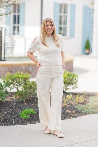 A full-length front view of a woman wearing the Sandbar Stroll Wide-Leg Pants, a pair of creamy ivory high-waisted trousers that radiate "old money" elegance. The pants feature a dramatic wide-leg silhouette and sophisticated vertical front seams designed to create an elongating, statuesque profile. Styled for a pristine monochromatic look with a matching cream short-sleeve knit and neutral Mary Jane flats against a bright, architectural outdoor backdrop.