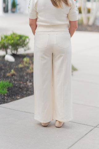 A full-length back view of the Sandbar Stroll Wide-Leg Pants emphasizing the clean, uninterrupted lines of the high-fashion wide-leg cut. The ivory "Sandbar" hue provides a crisp, sun-bleached aesthetic that pairs seamlessly with the tucked-in cream knit top. The view highlights the flattering fit at the natural waist and the generous, flowing hemline that accommodates various footwear styles.