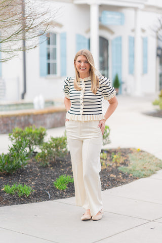 A full-length view of a woman wearing the Madeline Ruffle Knit, a cream and black horizontal striped short-sleeve cardigan. The top features a ruffled round neckline, a button-down front with delicate ruffle trim along the placket, and elbow-length puff sleeves with ruffled cuffs. It is styled with high-waisted cream wide-leg trousers and neutral pointed-toe flats, set against a bright, upscale outdoor shopping backdrop with light blue shutters and a fountain.