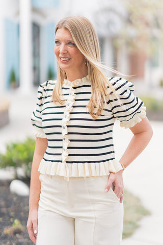 A portrait view highlighting the intricate textures of the Madeline Ruffle Knit. This shot showcases the soft-touch striped fabric and the feminine ruffle detailing on the collar and button-front closure. The elbow-length puff sleeves add architectural volume, while the wide ruffled hemline cinches the waist. The model is accessorized with classic gold hoop earrings and a gold bracelet, embodying a "Parisian chic" aesthetic.