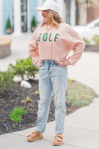 A woman posing outdoors in the Judy Blue Driftwood Boyfriend Jeans, featuring a relaxed, light-wash blue denim. She is wearing the mid-rise jeans with a peach-colored "GOLF" sweatshirt, a white baseball cap, and tan platform cork sandals. The jeans exhibit a roomy fit through the hips and thighs, intentionally designed for a laid-back, "effortless cool" aesthetic.