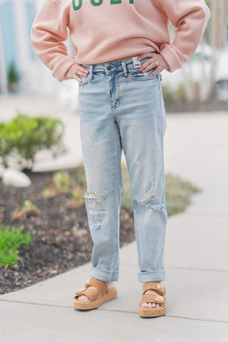 A close-up view of the Judy Blue Driftwood Boyfriend Jeans highlighting the sun-faded "Driftwood" wash and mid-rise waistband. The image showcases the signature artisanal repair stitching over distressed patches on the thighs, providing a vintage, mended look. The functional 5-pocket construction and classic button-zip fly are visible, paired with a tucked-in peach sweatshirt.