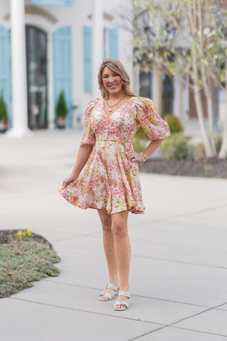 A full-length front view of a woman wearing the Sun-Drenched Petal Mini Dress, a romantic ivory mini dress with a sun-drenched botanical print in shades of pink, yellow, and peach. The dress features a flattering fixed V-neckline, half-length puff sleeves with elasticized cuffs, and a flirty flared skirt. It is styled with cream-colored block-heel sandals and a chunky gold pearl necklace, posed in a bright outdoor plaza.