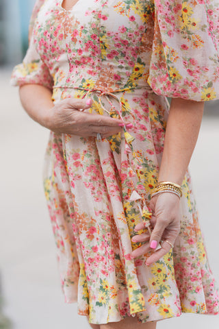 A detailed close-up of the Sun-Drenched Petal Mini Dress highlighting the delicate floral pattern and the slim, matching fabric tie-belt at the waist. The shot showcases the airy, lightweight woven fabric and the intricate gathered detailing on the puff sleeves. The model’s hands are shown adjusting the waist tie, emphasizing the customizable, figure-flattering fit.