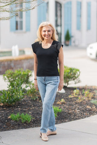 A woman smiling while posing in the Judy Blue Echo Wash Denim, a pair of slim-fit mid-rise jeans with a medium-toned vintage indigo wash. She is wearing a tucked-in black scoop-neck top and nude tassel loafers. The jeans feature an integrated, discreet cellphone pocket and authentic whiskering at the hips, providing a sleek and functional silhouette.