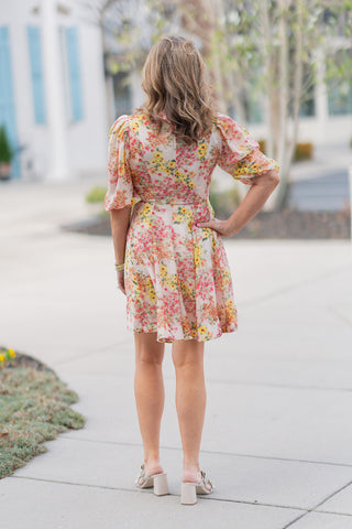 A full-length back view of the Sun-Drenched Petal Mini Dress emphasizing its ethereal movement and feminine silhouette. The botanical print covers the back and sleeves uniformly, while the flared mini skirt highlights the youthful and playful swing of the fabric. Paired with neutral block heels, this angle demonstrates the dress’s elegant drape and high-end boutique fit.