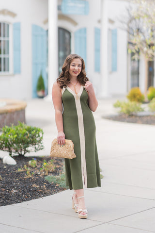 A full-length front view of a woman posing in the Annalise Colorblock Midi Dress, a sophisticated olive green ribbed knit midi dress. The design features a striking vertical cream center stripe that runs the full length of the column silhouette, creating a visually elongating effect. The dress is styled with a deep V-neckline framed by contrast cream banding and slim fixed straps. The model is accessorized with cream platform espadrille sandals and a textured straw lace clutch, set against a bright, archite