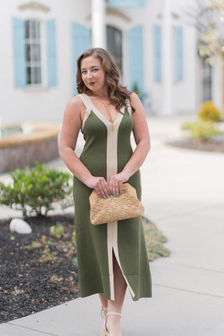 A side profile view of the Annalise Colorblock Midi Dress highlighting the premium, mid-weight ribbed knit fabric and the figure-skimming fit. The image showcases how the contrast cream trim transitions seamlessly from the deep V-neckline into the straps, emphasizing the clean lines and "quiet luxury" aesthetic. The model holds a tan lace-patterned clutch with a wooden frame, highlighting the organic olive and cream color palette.