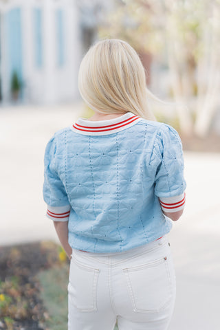 The back view of the Court Club Polo emphasizing its tailored fit and textured breathability. The delicate pointelle knit covers the back and puff sleeves, while the red-striped white collar and hem provide a structured frame. Paired with white trousers, this angle showcases the slightly blouson effect created by the wide ribbed waistband.