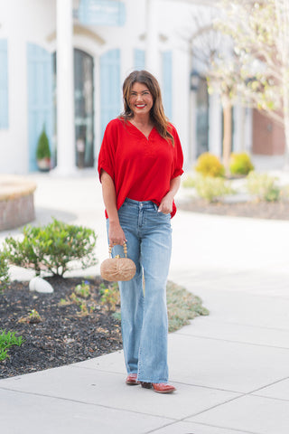 A woman smiling outdoors wearing the Judy Blue V-Line Vault Jeans, a pair of medium-wash baggy streetwear jeans. She has styled the high-rise, V-front denim with a vibrant red short-sleeved blouse, brown western-style boots, and a small straw clutch with a beaded handle. The jeans feature a relaxed silhouette and a subtle raw-edge hem.