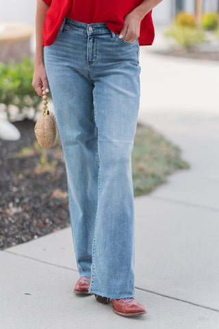 A detailed front view of the Judy Blue V-Line Vault Jeans highlighting the unique V-shaped asymmetrical waistband and medium-wash finish with natural fading. The image showcases the relaxed, spacious fit of the streetwear-inspired design and the clean raw-edge hem stacked over reddish-brown leather boots.
