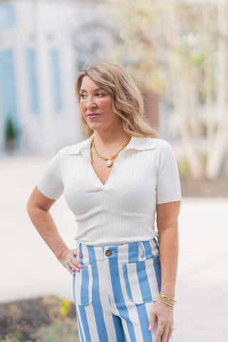 A full-length front view of a woman posing in the Pier Side Polo, a bright white ribbed knit top. The polo features a classic fold-over collar with a buttonless V-placket and a slim, body-contouring fit. It is styled for a "coastal-chic" look, tucked into high-waisted light blue and white vertical striped wide-leg trousers and paired with neutral block-heel sandals. The model is standing in a bright outdoor plaza with light blue shutters in the background.