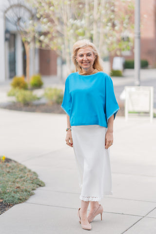 A full-length front view of a woman wearing the Willow Whisper Midi Skirt, a pristine ivory midi skirt that epitomizes "quiet luxury". The skirt features a flattering high-rise waist and a subtle A-line flare, styled with a vibrant turquoise poncho-style top with white whipstitch detailing. The elegant silhouette is finished with a delicate eyelet lace insert and a scalloped ruffle hem, paired with nude pointed-toe pumps in an upscale outdoor setting.