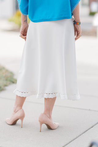 A close-up back view of the Willow Whisper Midi Skirt emphasizing the clean, uninterrupted fit and graceful drape of the fluid fabric. The ivory hue acts as a luminous blank canvas, highlighting the sophisticated profile and the detailed eyelet and ruffled hemline from behind. Styled with neutral heels, this angle demonstrates the skirt's statuesque and visually lengthening effect.