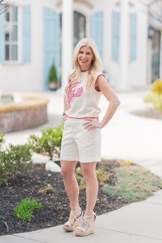A full-length view of a woman posing in the Cape Cod Catch Knit, a whimsical cream sweater vest featuring a bold, oversized lobster graphic in red gingham. The vest is designed with a sleeveless silhouette, a round neckline, and a slightly cropped hem, all finished with intricate red crochet-style whipstitching. Styled for a "coastal chic" look, it is paired with high-waisted cream tailored shorts and tan platform wedge sandals against a bright, architectural outdoor backdrop.