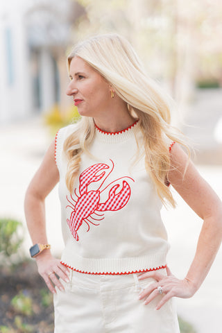 A portrait view highlighting the artisanal details of the Cape Cod Catch Knit. The shot showcases the vibrant red and white gingham pattern of the centered lobster motif and the delicate red contrast stitching along the crew neck and armholes. The smooth, premium cream knit fabric offers a refined, boutique-exclusive feel, while the model’s blonde hair and classic gold earrings complement the preppy, nautical aesthetic.