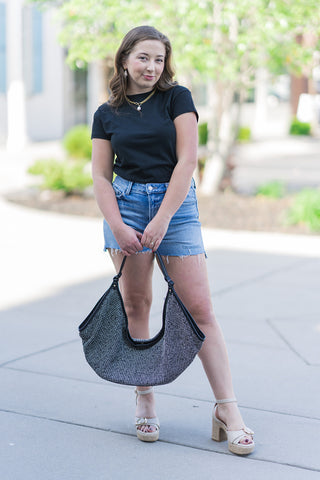 A woman poses outdoors wearing the Boardwalk Bound Shorts, a pair of high-rise denim cut-offs in a medium-light wash. She styles them with a classic black crew neck tee, a large shimmering silver-and-black tote bag, and cream-colored platform espadrille sandals. The shorts feature a trendy raw side step hem that elongates the leg, creating a perfect fashion-forward summer look.