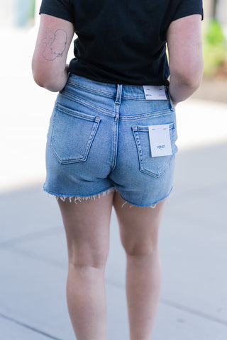 The back view of the Boardwalk Bound Shorts emphasizes the traditional five-pocket design and the Vervet brand tag on the waistband. The high-rise fit and raw, unfinished hem are clearly visible, illustrating the edgy, contemporary flair of the garment. The image highlights the shape retention of the premium stretch denim from behind as the woman poses.