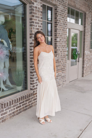 A three-quarter view of the Gilded Garden Gown, capturing the light-reflecting qualities of the sequined floral embroidery. The model poses near a storefront window, showing how the drop-waist silhouette and tiered hemline create a modern, high-fashion elongated profile. The soft cream hue and intricate textures make it an ideal choice for bridal events or upscale garden parties.