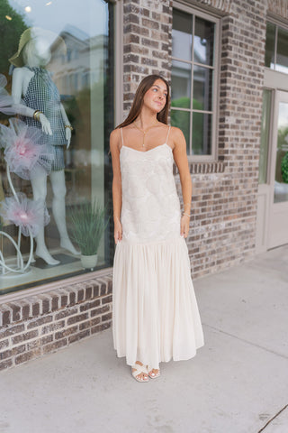 A woman stands outside a brick boutique wearing the Gilded Garden Gown, a luxurious cream-colored maxi dress. The gown features a straight neckline with delicate spaghetti straps and an intricate bodice adorned with oversized tonal floral appliqués, sequins, and beadwork. The design transitions into a low drop-waist seam that gives way to a voluminous, pleated tiered maxi skirt, styled with gold jewelry and ivory woven slide heels.