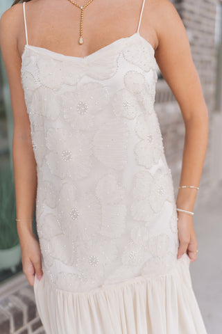 A detailed close-up of the Gilded Garden Gown highlighting the artisanal craftsmanship of the bodice. Large, shimmering floral embroidery and 3D floral appliqués are accented with delicate pearl-like beads and sequins for a multidimensional texture. The model wears a gold drop-pendant necklace and layered pearl bracelets, emphasizing the dress’s elegant V-shaped bust detail and "quiet luxury" aesthetic.