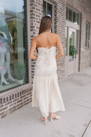 Rear view of the Gilded Garden Gown showcasing the feminine back details. The cream bodice features a thin tie-back closure and a subtle ruffle detail along the spine, maintaining a sleek, adjustable fit. The low drop-waist transition leads into a lightweight, flowing skirt that pools elegantly at the floor, highlighting the dress's architectural silhouette and graceful movement.