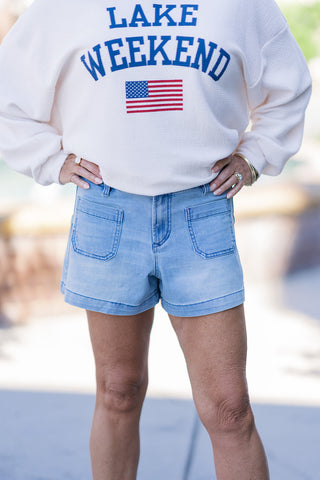 A close-up front view highlighting the detailed design of the Dear John Carlisle Eclipse Shorts. Sitting comfortably at a mid-rise fit, the light-medium blue denim features prominent dual front patch pockets that offer a retro, utilitarian flair. The structured waistband includes classic belt loops and a secure zip-fly closure, transitioning down to a thick, polished clean hemline. The model is wearing a white sweatshirt with a "Lake Weekend" graphic and an American flag.