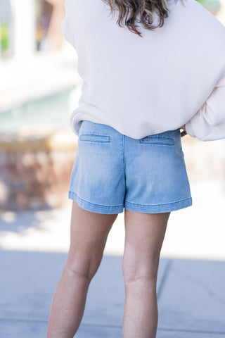 Back view of the Dear John Carlisle Eclipse Shorts, showcasing the clean and pocket-free rear silhouette of these premium denim bottoms. The light-to-medium indigo wash fabric drapes smoothly into a relaxed leg opening finished with a structured, un-distressed clean hem. The model styles the shorts casually with a cream-colored, oversized waffle-knit pullover that rests at the mid-rise waistband.