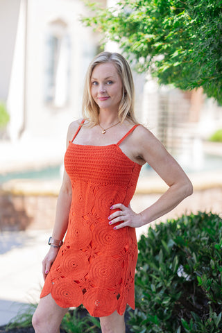 A portrait view of the Sicilian Solstice Mini Dress focusing on the textured detailing of the bodice. The dress features delicate spaghetti straps and a straight-across square neckline with a densely crocheted upper band, which elegantly gives way to large, open-work abstract circle-and-starburst crochet motifs. The blonde woman poses with one hand on her hip, accessorizing the summery tangerine look with a simple gold heart pendant necklace.