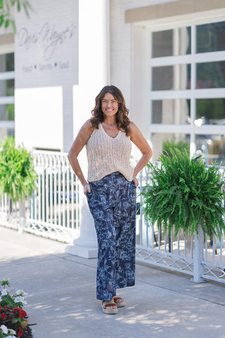 A full-length outdoor portrait of a smiling woman posing to demonstrate the lifestyle styling of the Dear John Ulyssa Top. The sleeveless tank features a premium open-weave crochet texture in a soft neutral cream shade with a classic V-neckline. She styles the fluid, relaxed knit top with navy blue wide-leg trousers adorned with a light blue floral pattern and pairs the look with tan leather platform espadrille sandals.