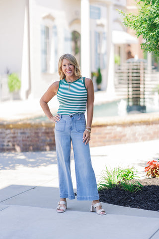Full-length outdoor portrait of a smiling woman posing to display the complete lifestyle outfit featuring the Dear John Audrey Eclipse Jeans. She stands with one hand on her hip, pairing the high-rise, cropped wide-leg denim pants with a tucked-in green and white horizontally striped sleeveless top. The casual, sun-kissed boutique ensemble is accessorized with gold jewelry and cream block-heeled sandals, set against a blurred resort-style courtyard backdrop.