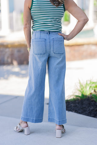 Rear view of the lower body demonstrating the fit and tailoring of the Dear John Audrey Eclipse Jeans. The high-waisted denim pants highlight a supportive fit with clean, vertical dart detailing above two deep, functional back patch pockets. The light-wash stretch fabric falls into a relaxed, wide-leg silhouette that stops just above the ankle to showcase off-white block-heel mules.